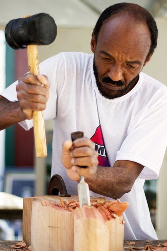 Emerging Artist man holding chisel and mallet carving a block of wood on a table